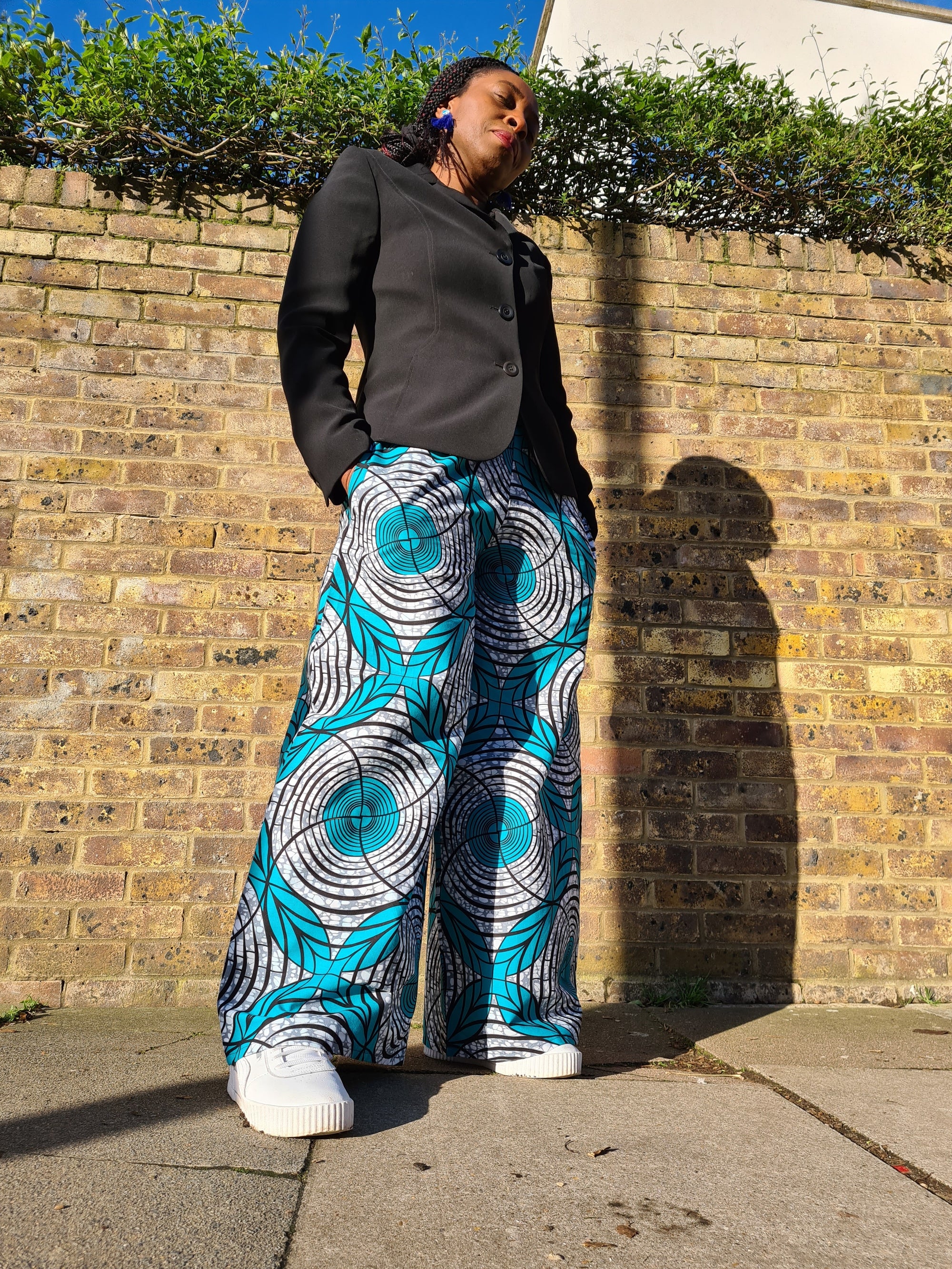 Lena King Black woman standing in front of a brick wall wearing a pair of teal and grey trousers, a black suit jacket and white trainers.  She has her hands in her pocket and is wearing a pair of blue earrings.