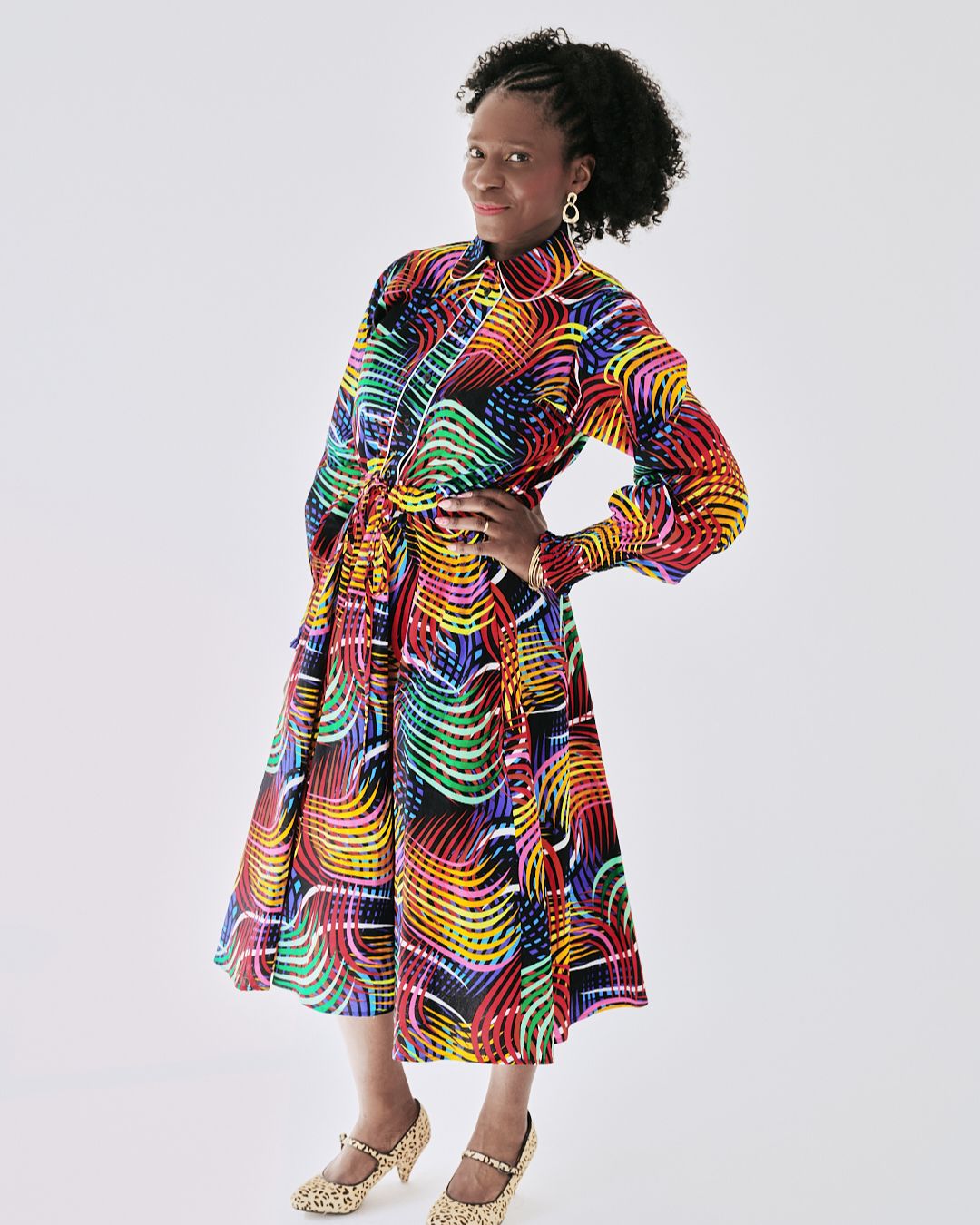 A model wears a vibrant, multicolor swirl-patterned shirt dress. The midi-length dress features long sleeves, a collared neckline, a flared skirt, and a waist tie. The swirling rainbow hues pop against a black background.