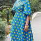 A woman radiantly posing in the long blue print puff sleeve dress paired with blue trainers in a park setting.