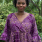 A headshot of a woman happily wearing the purple print ruffle dress in a park setting.