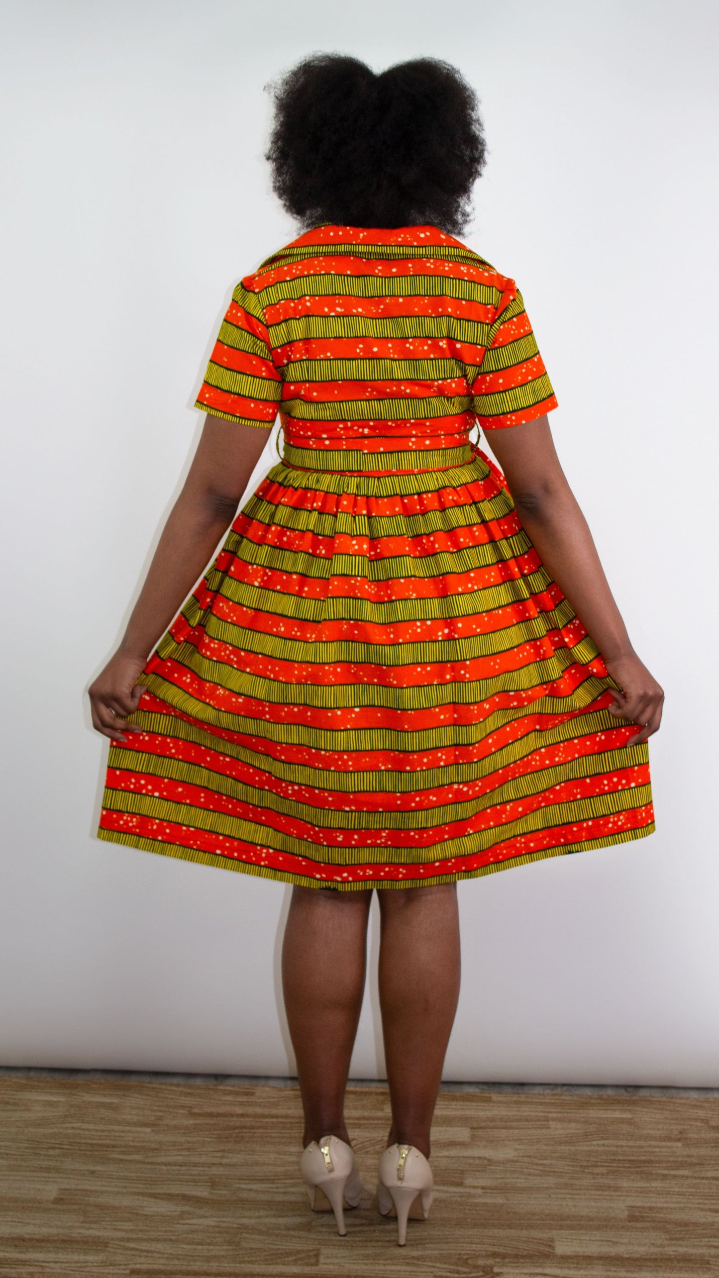 A model elegantly reveals the back of a short orange-yellow striped print dress, gracefully holding and lifting the dress to showcase its charming details. 