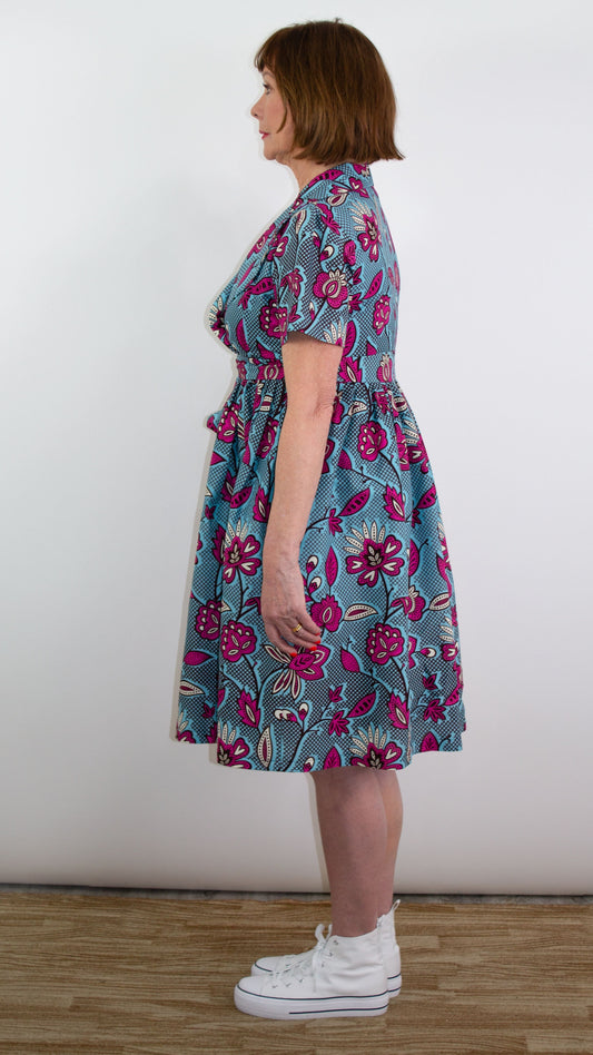 A side view capturing a model sstanding in a blue dress with a pink floral print, elegantly showcasing the silhouette of the dress, paired with white trainers.