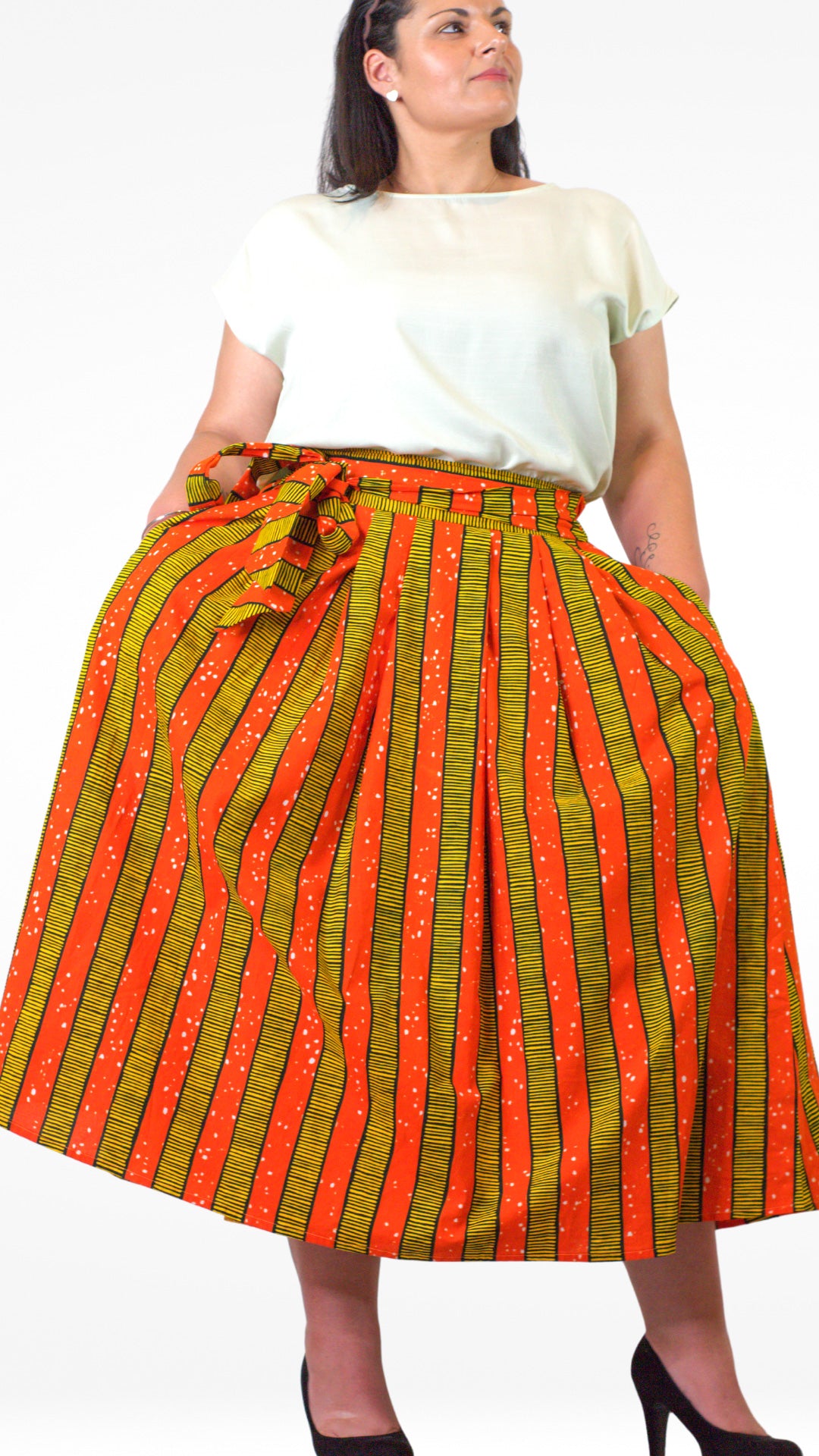 a person in a orange yellow striped skirt wearing a white top and black heels and posing with arms on waist standing confidently