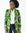 A model showcases the tailored Ankara blazer, its green floral pattern vibrant against the pink and white background. Paired with a crisp white shirt and dark jeans, the image emphasizes the blazer's bold print and fitted silhouette.