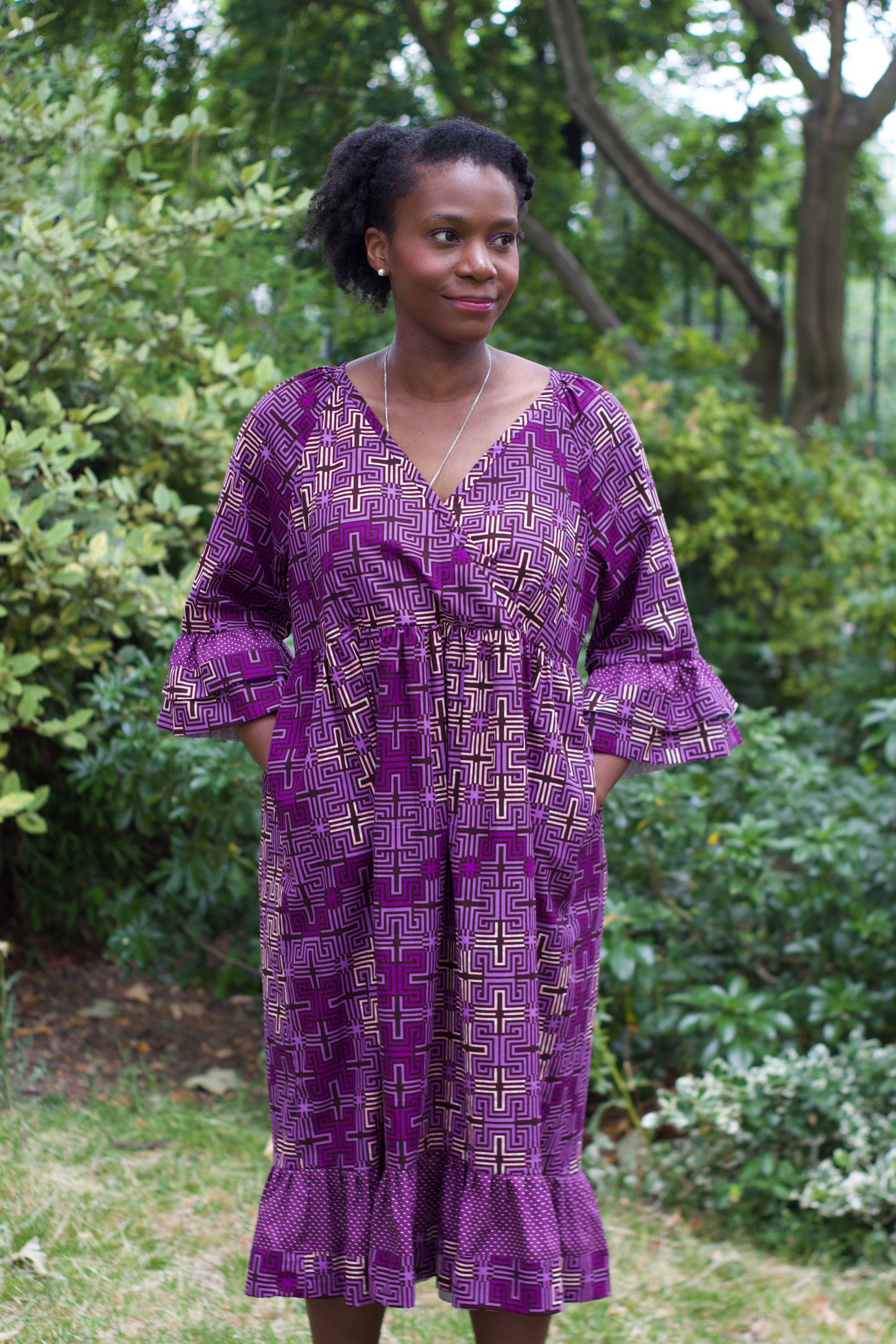 A woman wearing the purple Dawn African Print Ruffle Dress by Dovetailed London in a park setting, casually having placed hands in pockets, exuding comfortability.