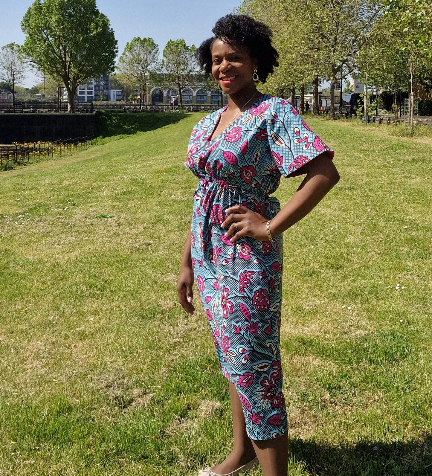 A woman posing confidently in a blue print pink elements kaftan style dress in a park setting, wearing pink ballet flats.