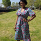 A woman strikes a pose in a stylish blue kaftan dress adorned with a pink floral print, set against a park. 