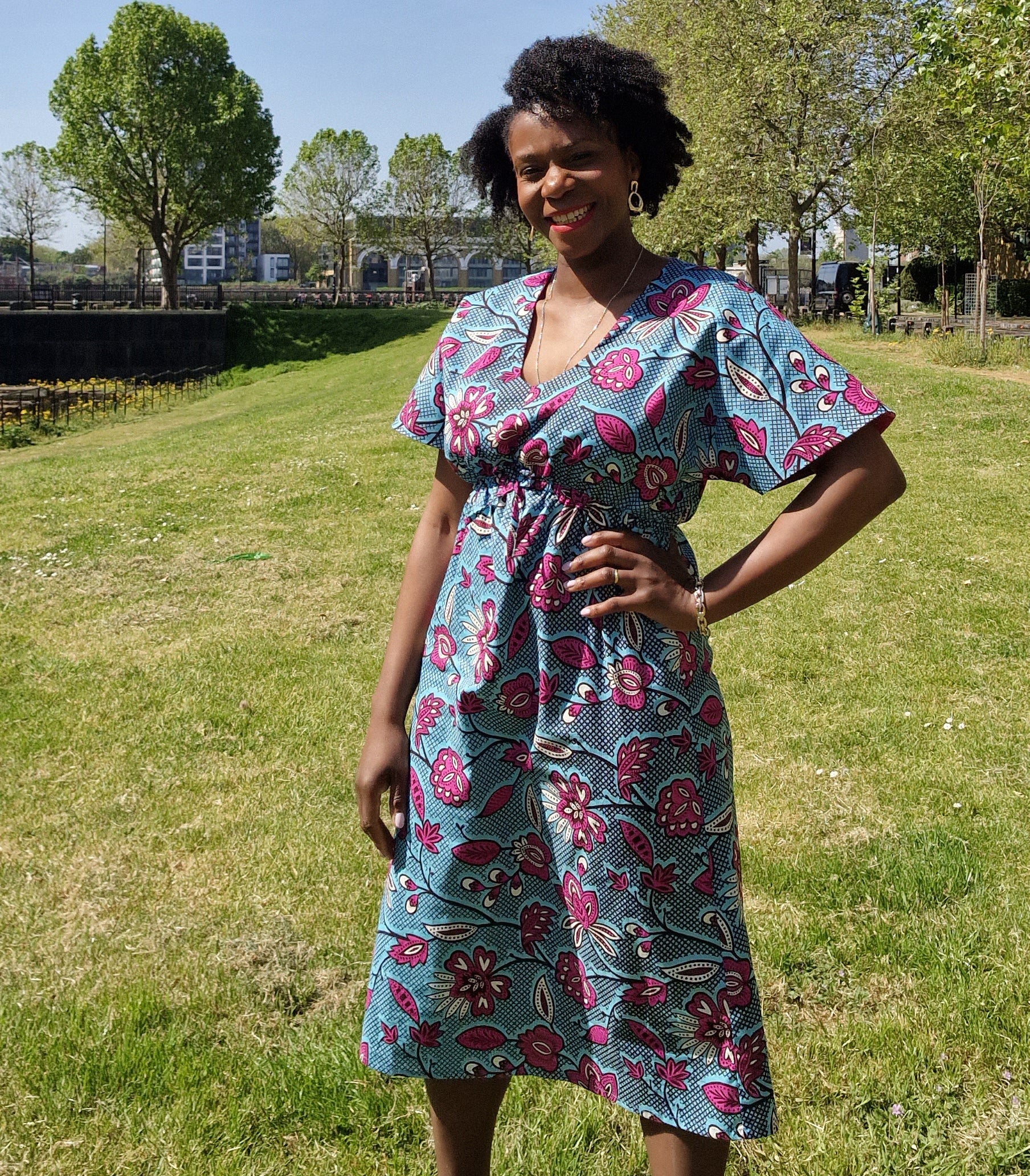 A woman strikes a pose in a stylish blue kaftan dress adorned with a pink floral print, set against a park. 