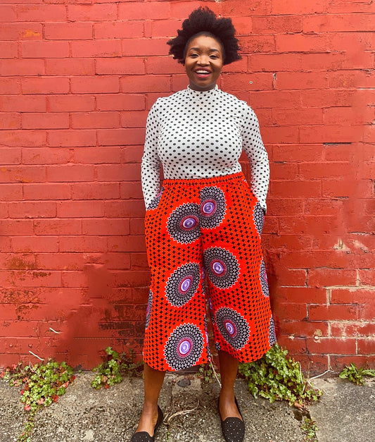 Black woman standing in front of a red brick wall wearing a pair of culottes made in African wax print fabric in red fabric with large circular motifs and a white top.