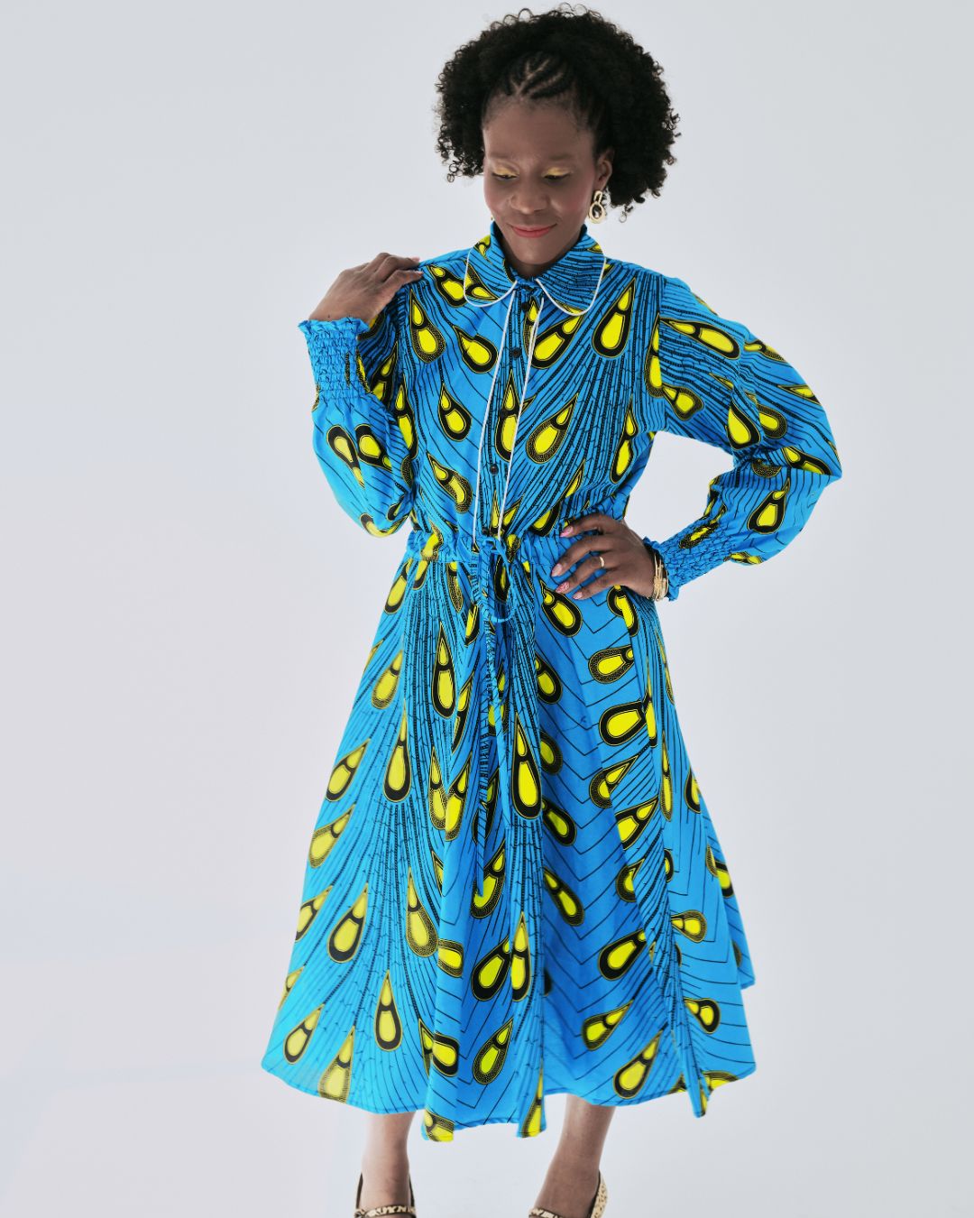 Model wearing a vibrant blue Ankara shirt dress with bold yellow teardrop pattern, full midi skirt, and long gathered sleeves. The dress features a classic collar and adjustable waist tie, styled for a flattering silhouette.