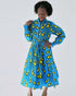 Model wearing a vibrant blue Ankara shirt dress with bold yellow teardrop pattern, full midi skirt, and long gathered sleeves. The dress features a classic collar and adjustable waist tie, styled for a flattering silhouette.