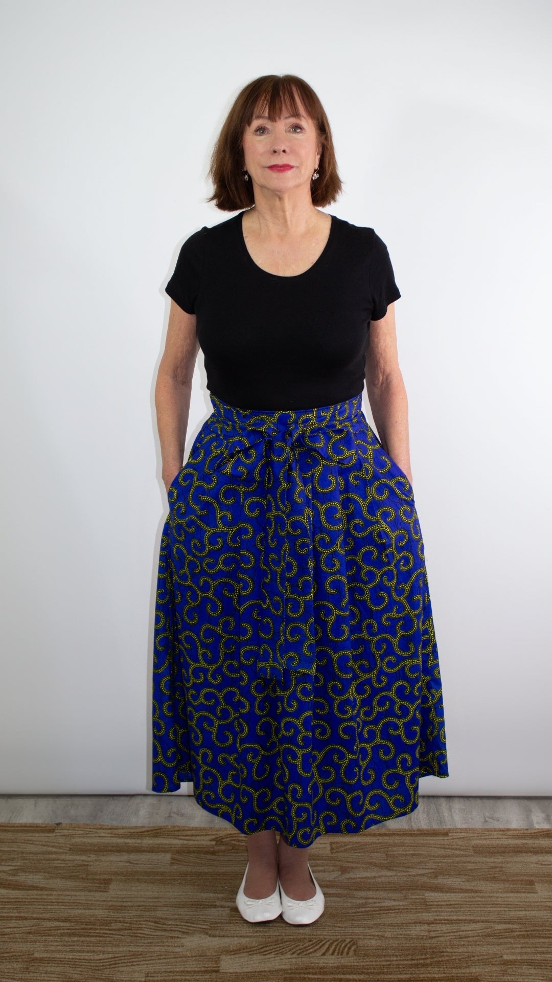 Model posing in a captivating blue swirly skirt adorned with golden elements paired with a chic black shirt. and white ballet flats.