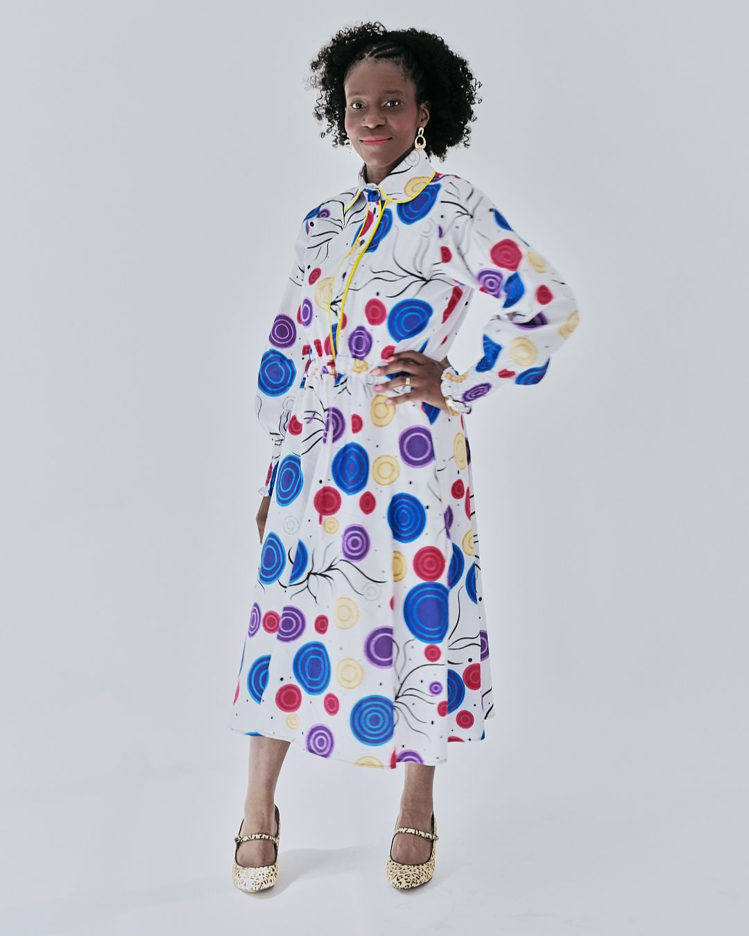 A woman poses in a white midi shirt dress adorned with a multicolored circular print. The dress features long puffed sleeves and a yellow piped button placket. One hand playfully holds the skirt.