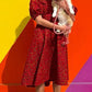 A person exudes happiness in a red puff-sleeve dress, paired with trainers and joyfully holding a corgi in front of a colourful wall background.
