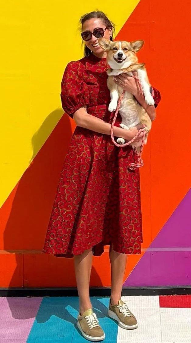 A person exudes happiness in a red puff-sleeve dress, paired with trainers and joyfully holding a corgi in front of a colourful wall background.