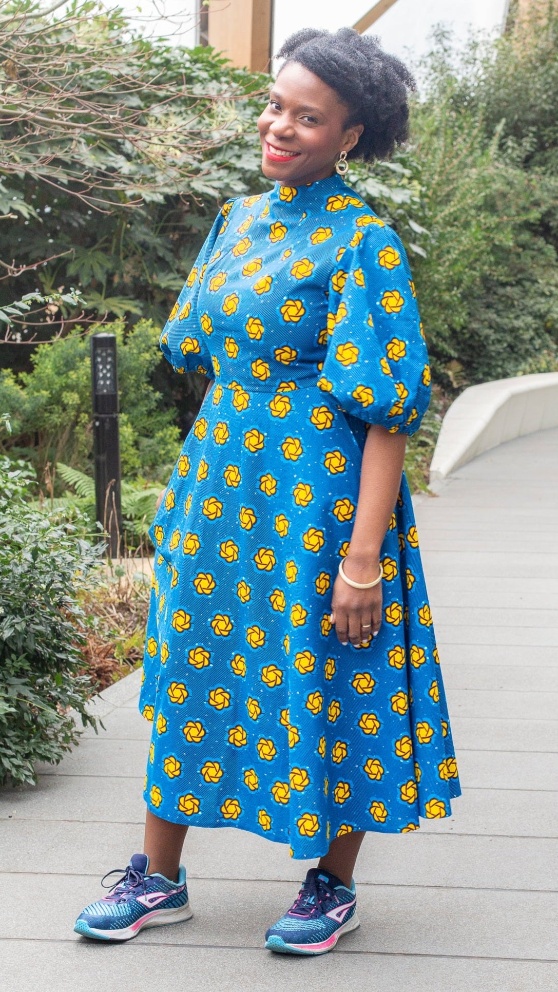 A woman radiantly posing in the long blue print puff sleeve dress paired with blue trainers in a park setting.