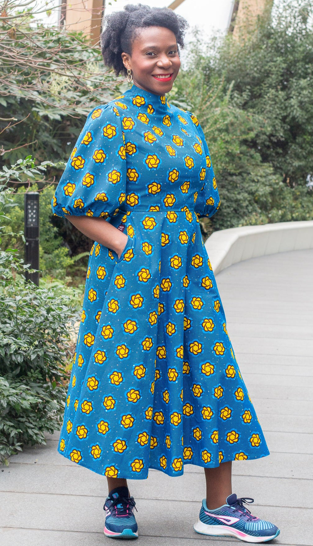 A woman posing with a smile in the long blue print puff sleeve dress in a park setting.
