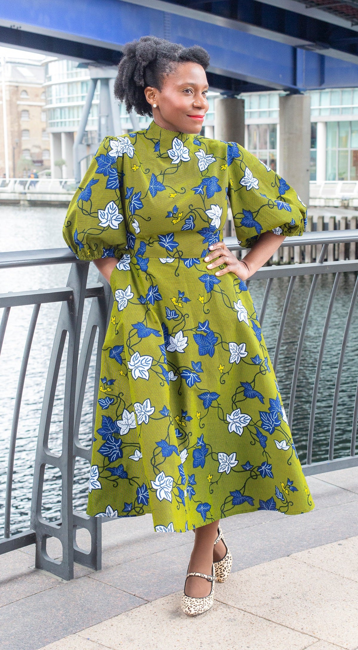 A woman confidently poses in a long khaki dress adorned with detailed blue and white leaves.