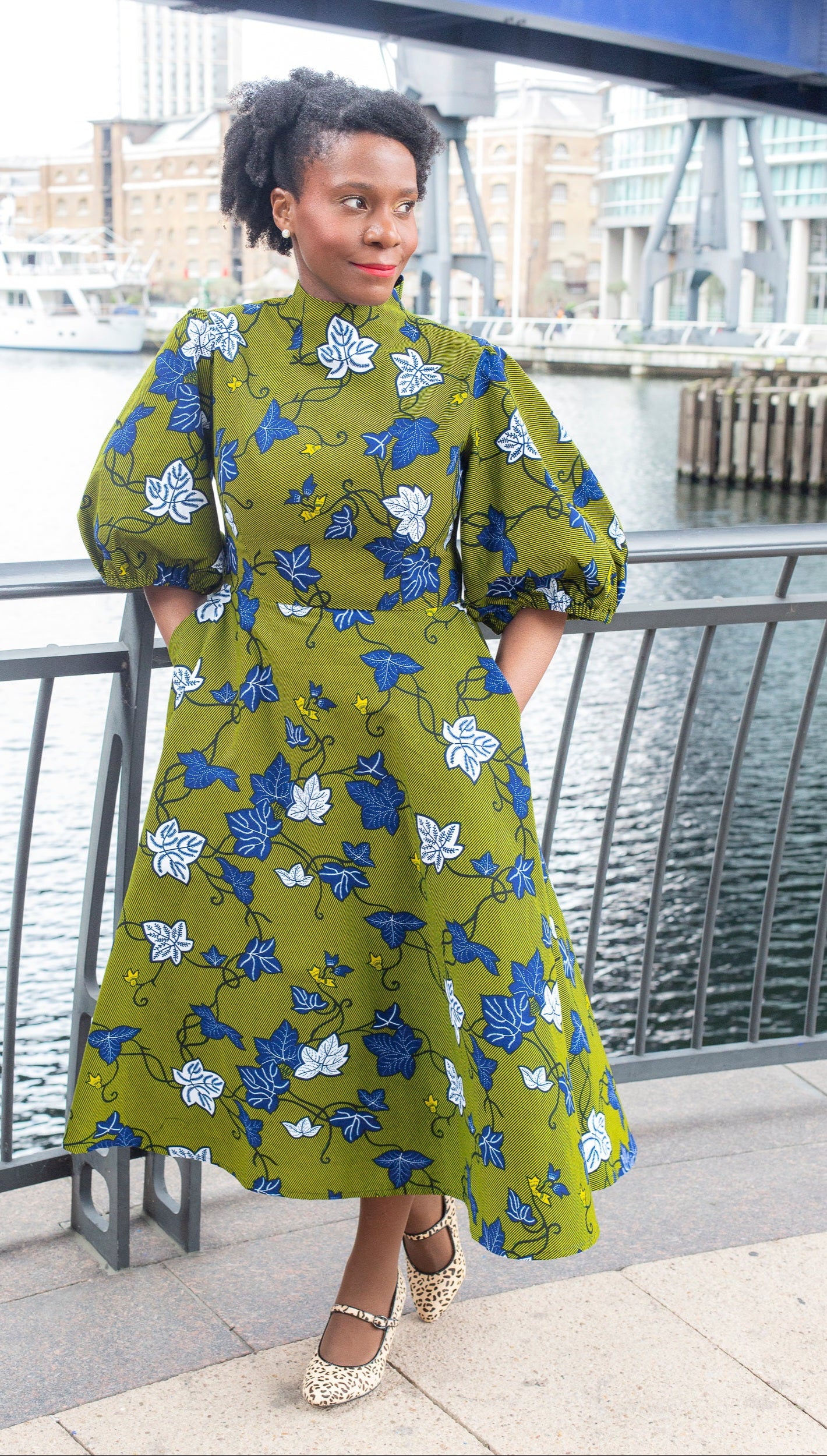 A woman posing on a bridge in a khaki dress adorned with detailed blue and white leaves.