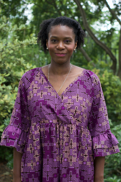 A headshot of a woman happily wearing the purple print ruffle dress in a park setting.