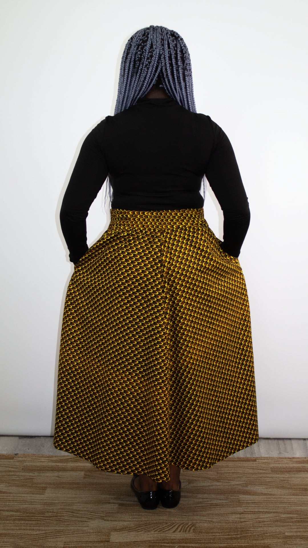 back of a model with vibrant blue hair displays a sleek black turtleneck coupled with a distinctive yellow and black skirt featuring an African print and accentuated by a trendy bow tie belt.