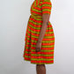 A model standing in a short orange-yellow striped print dress, featuring practical pockets and complemented by an elegant pair of beige heels. 