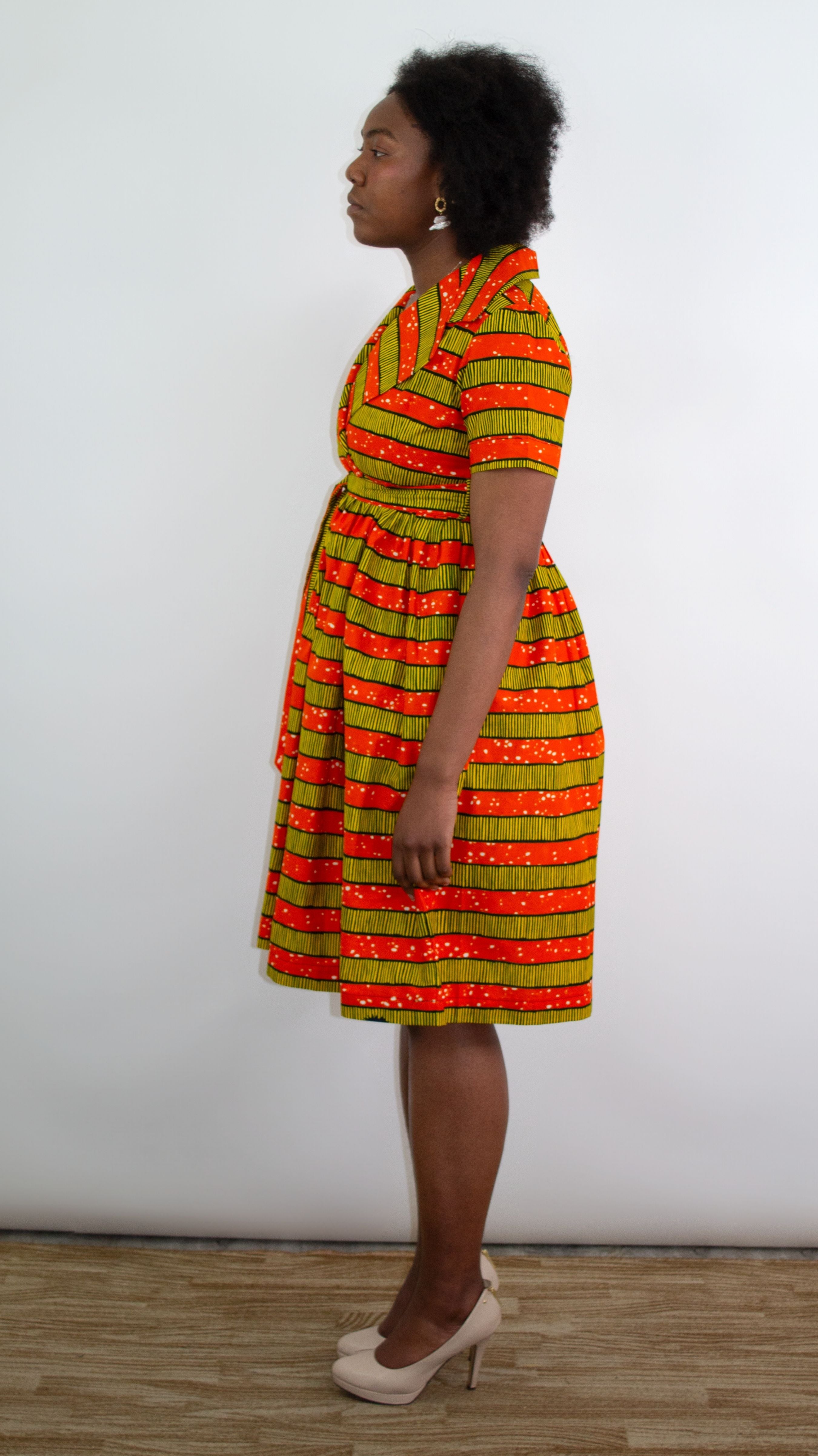 A model standing in a short orange-yellow striped print dress, featuring practical pockets and complemented by an elegant pair of beige heels. 