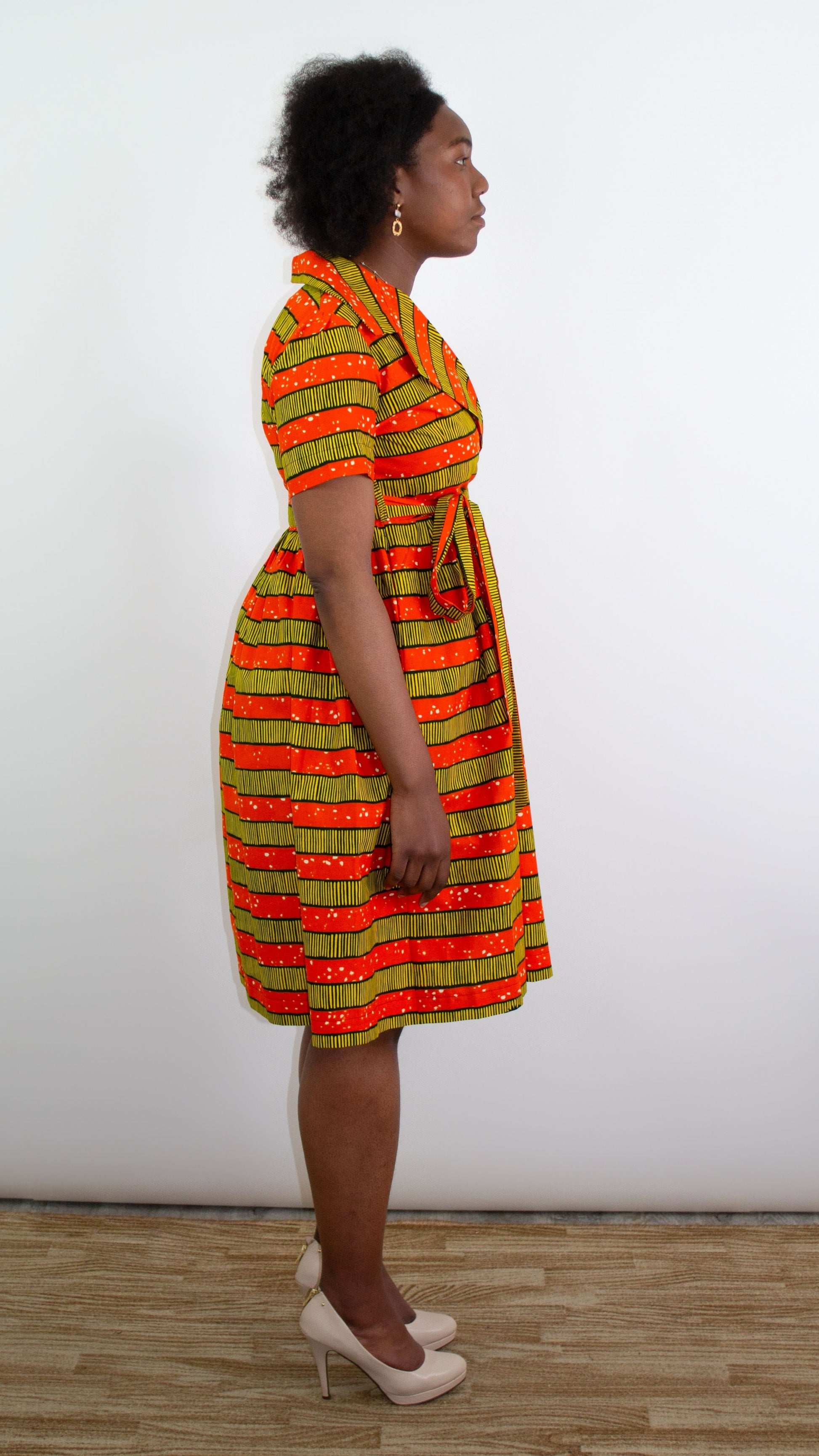 A model standing from the side reveals the tie bow belt of a short orange-yellow striped print dress.
