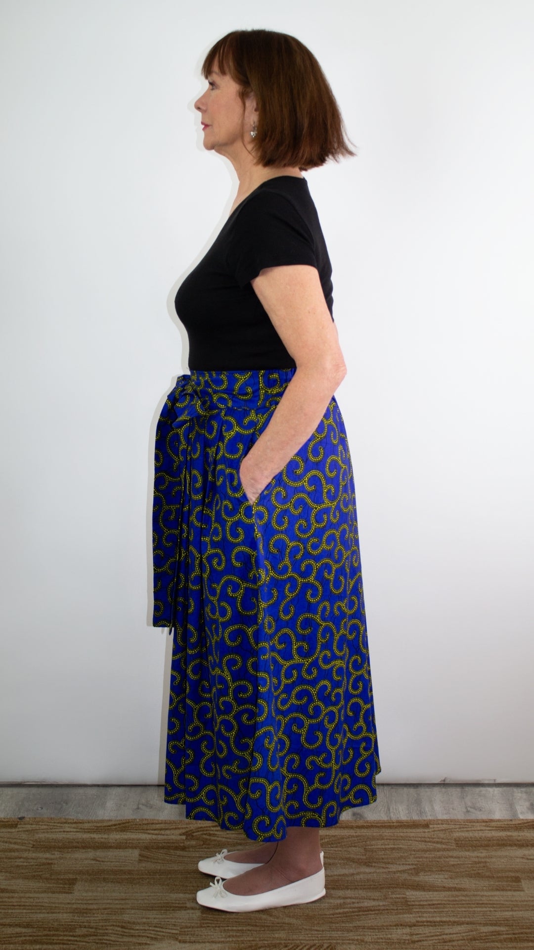 A side view of a model posing in a swirly blue print skirt with golden elements, highlighting the deep pockets and the beautiful silhouette of the skirt, emphasising both practicality and elegance.