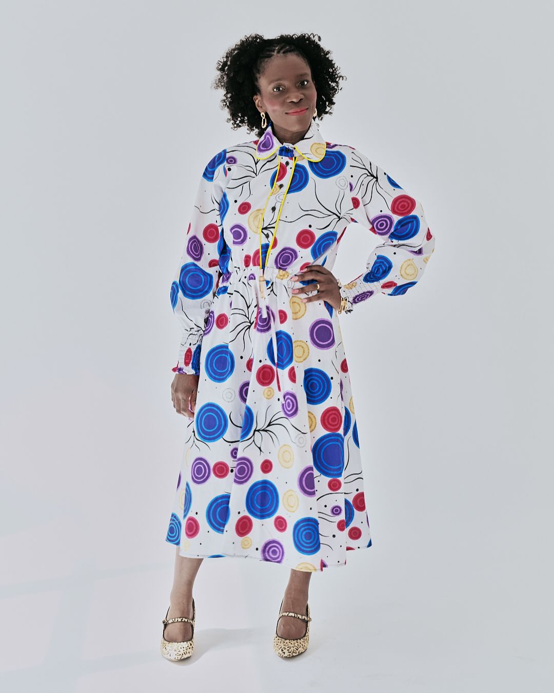 Alt Text: A woman poses in a white midi shirt dress adorned with a multicolored circular print. The dress features long puffed sleeves and a yellow piped button placket. One hand playfully holds the skirt.