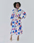 Alt Text: A woman poses in a white midi shirt dress adorned with a multicolored circular print. The dress features long puffed sleeves and a yellow piped button placket. One hand playfully holds the skirt.