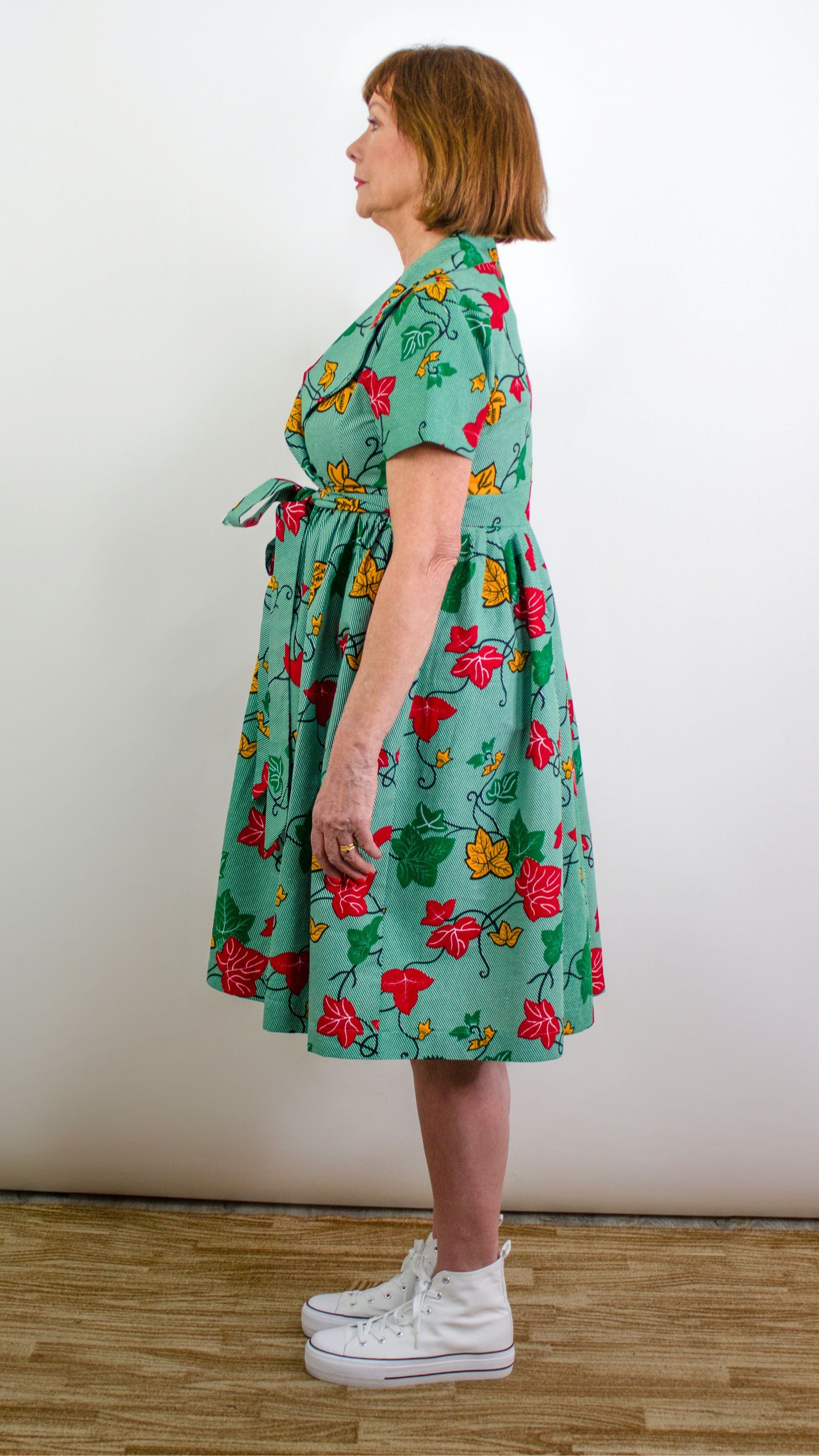 A side view of a model posing in a knee-length green print dress with leaf elements, highlighting the tie belt, adding a stylish accent to the ensemble and accentuating the natural flow of the dress.