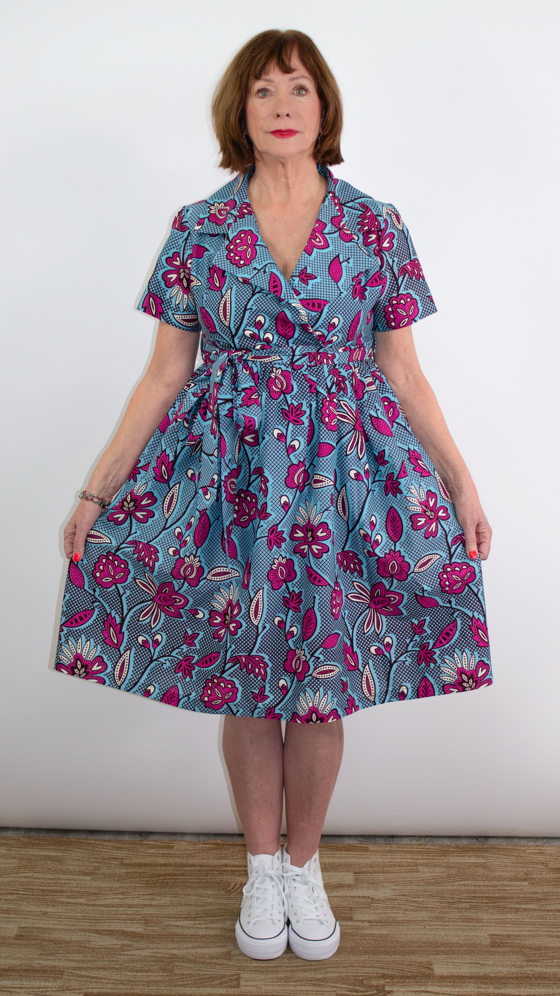 A model striking a pose in a blue dress with pink floral print, gracefully holding and lifting the dress. The lively floral patterns and wrap design, accentuated by this dynamic pose, create a stylish and flattering silhouette, capturing the essence of this eye-catching ensemble.
