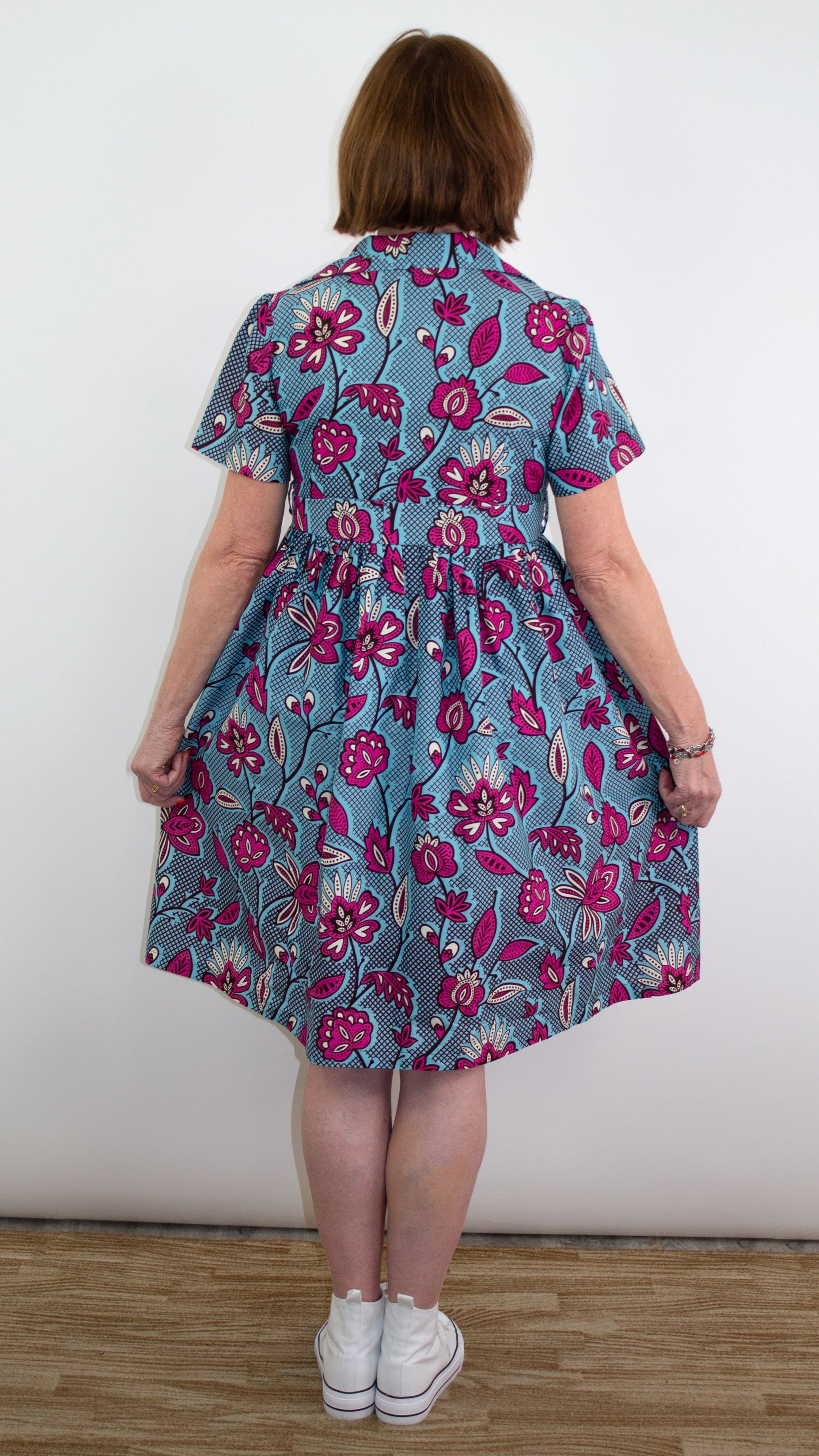 A model with short hair showcasing the back of a blue print dress with pink floral elements paired with white sneakers and a colourful bracelet.