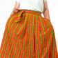 a person in a orange yellow striped skirt wearing a white top and black heels and posing with arms on waist standing confidently