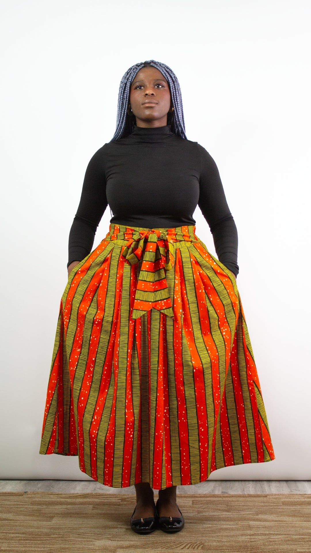 a full body portrait of a person posing in a long orange and yellow skirt with a tie belt, black top and black ballet shoes
