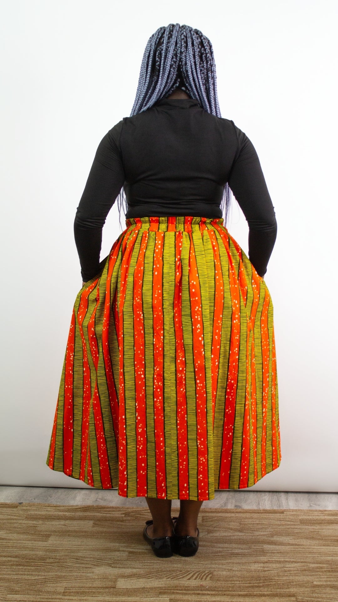 A full-body portrait capturing the back view of a person wearing a striking long orange and yellow skirt with a tie belt, complemented by a black top and stylish black ballet flats. 