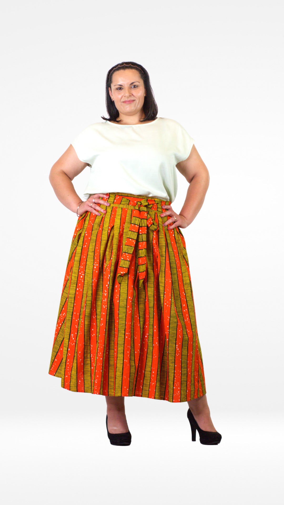 A pose of a model in an orange-yellow striped skirt paired with a crisp white top and stylish black heels. The person stands with arms confidently placed on the waist. 