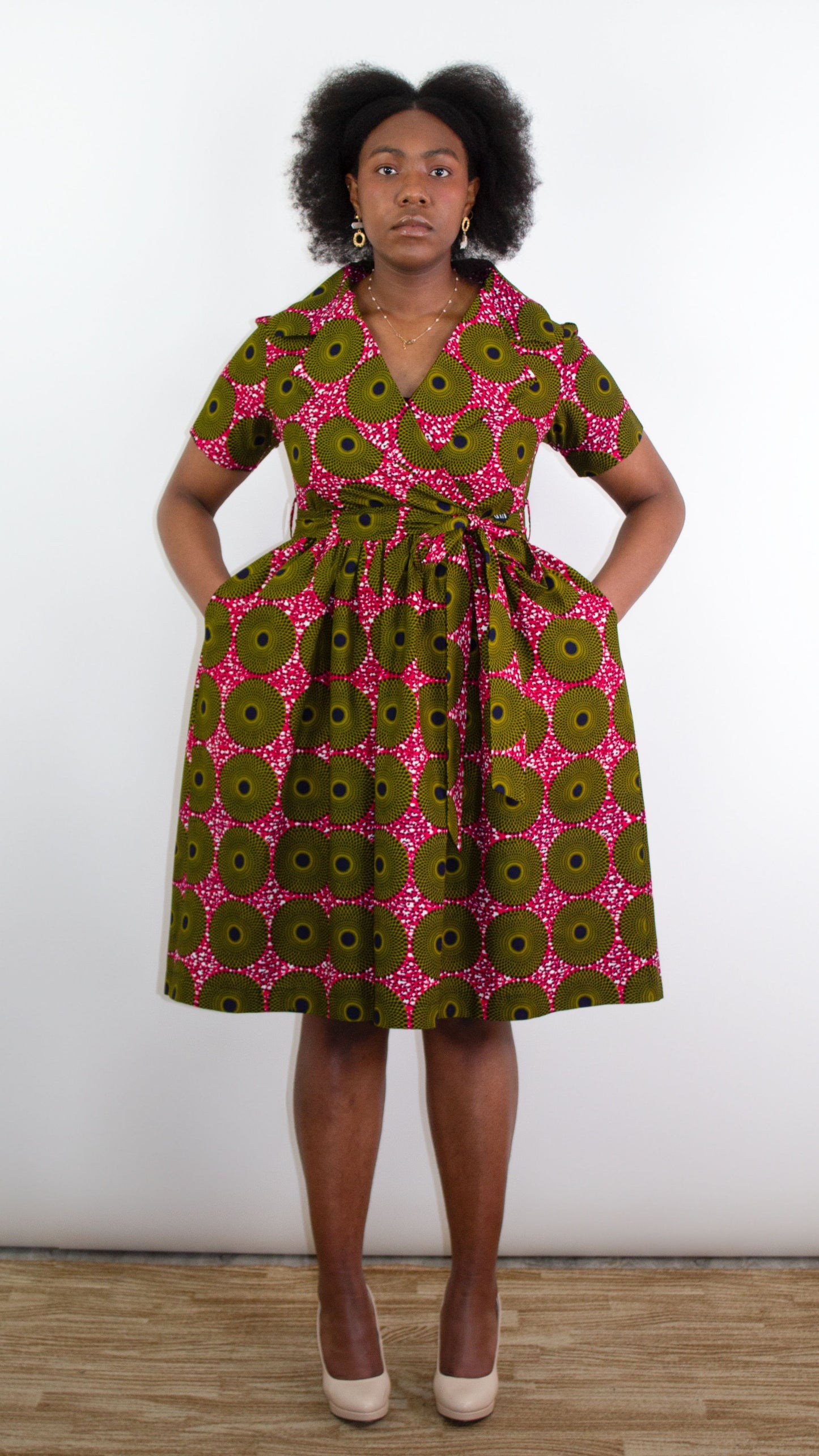 Model posing in the African Print 'Ripple Effect' Wrap Dress in pink fabric. Her hands are casually in the pockets and complemented by beige high heels.