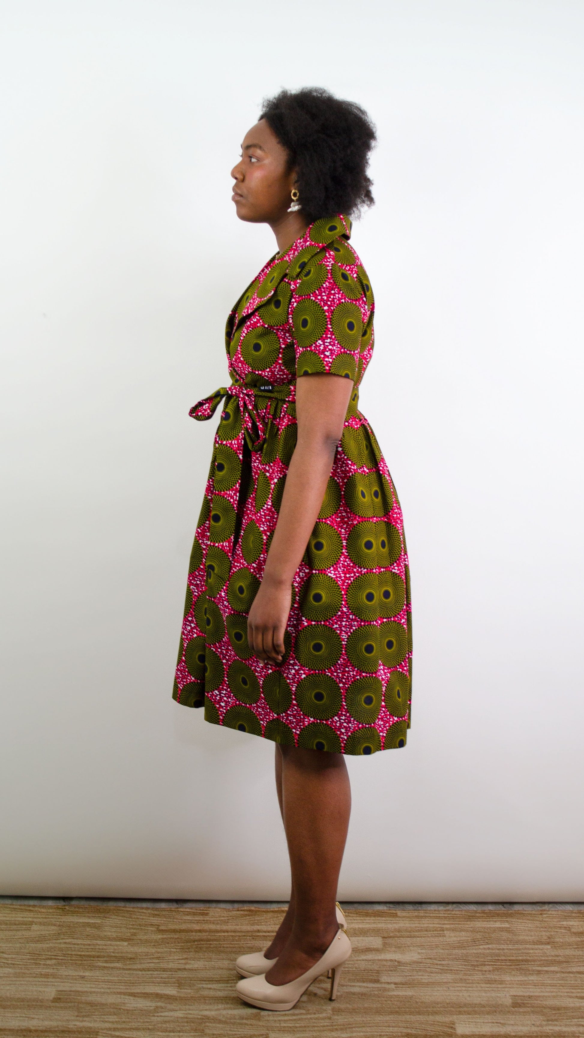 Side view portrait of a model posing in the pink fabric ripple effect wrap dress with hands over pockets, showcasing a bow tied belt.