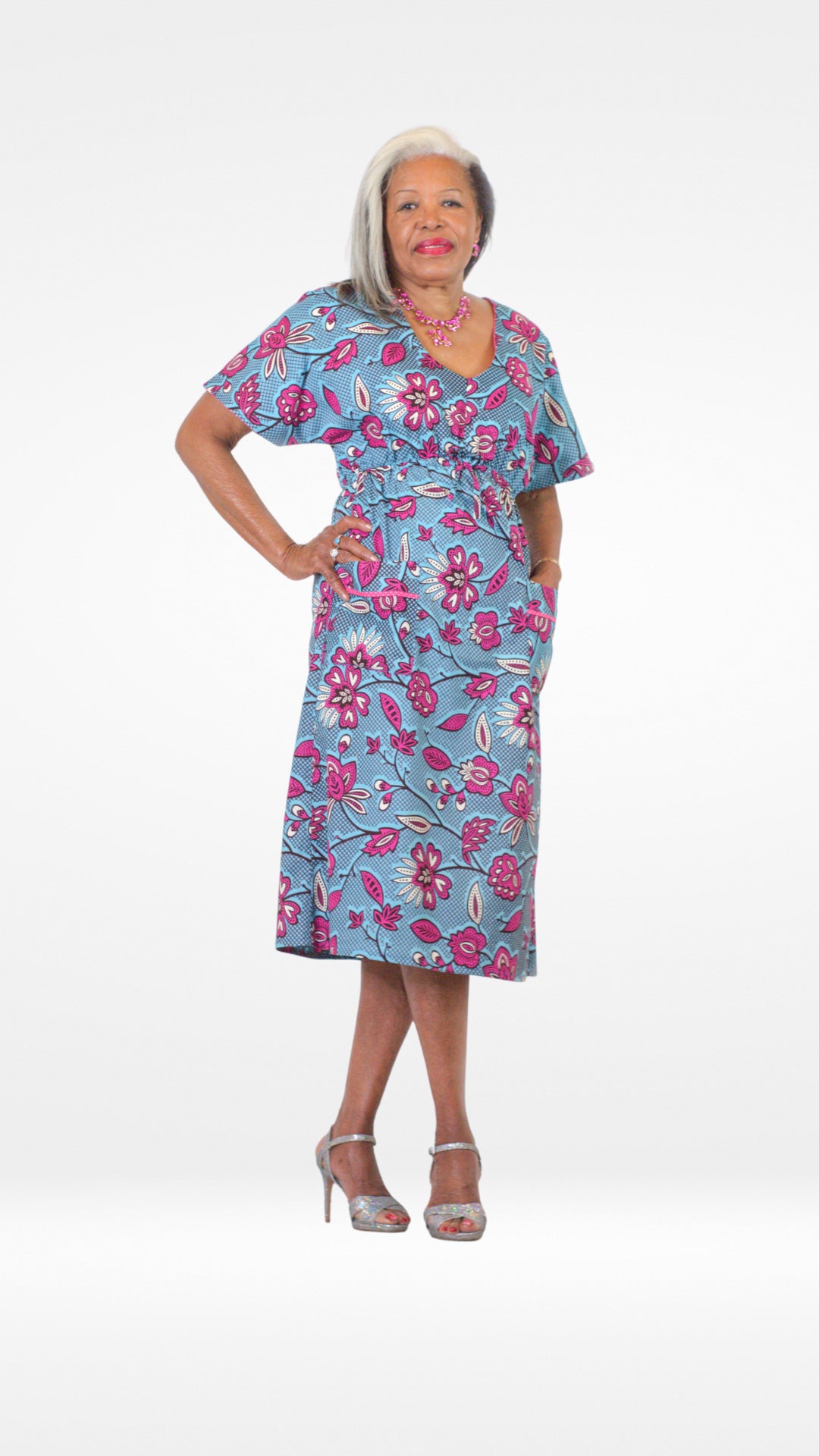 A model posing confidently in a blue kaftan dress with a pink floral print. The lively floral patterns, accentuated by this dynamic pose, create a stylish and flattering silhouette, capturing the essence of this eye-catching ensemble, paired with silver open toe high heels and a pink necklace.