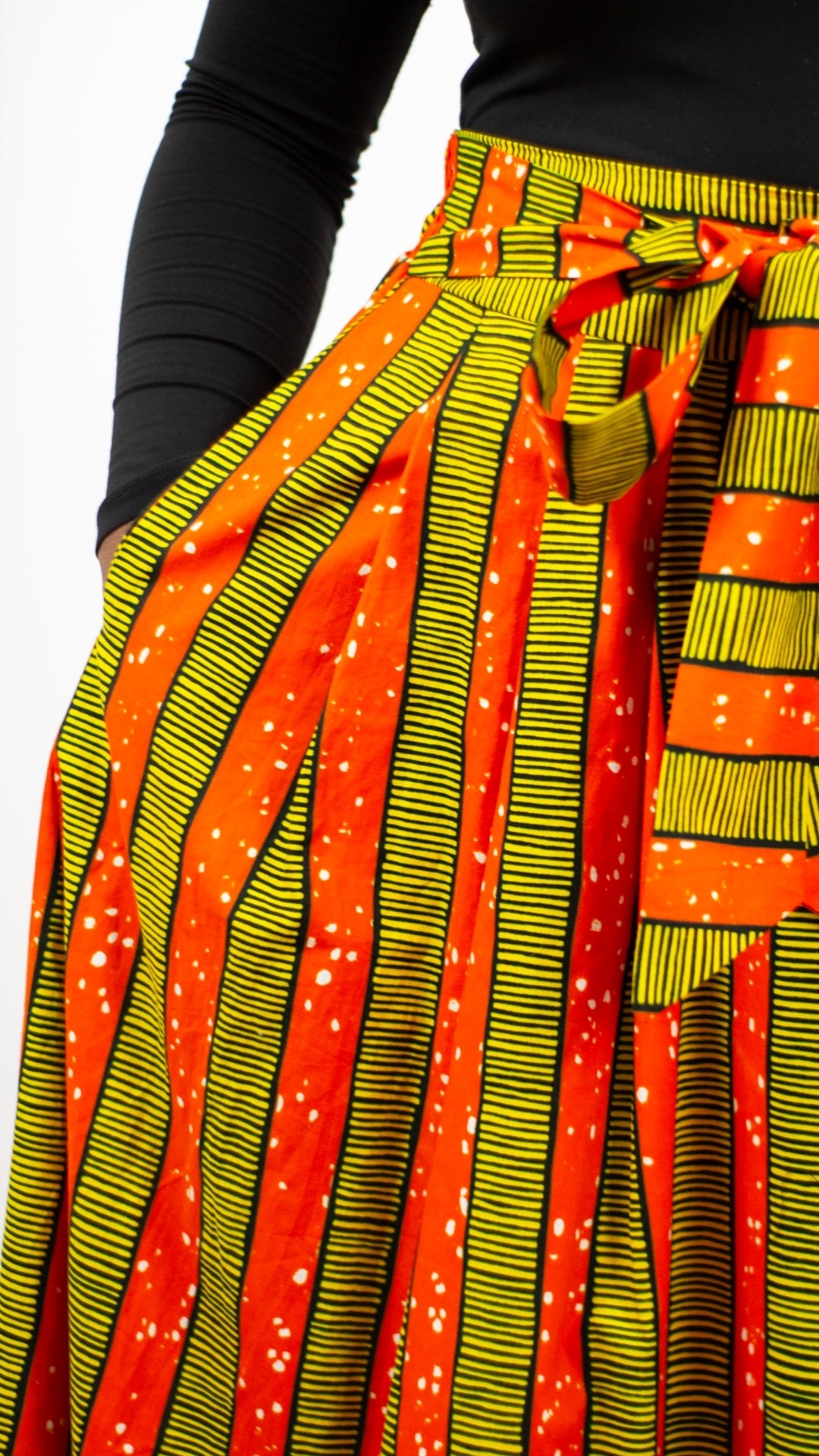 A person's hand in a yellow-orange striped skirt, emphasises the deep pockets and highlights the overall comfortability of the garment. 