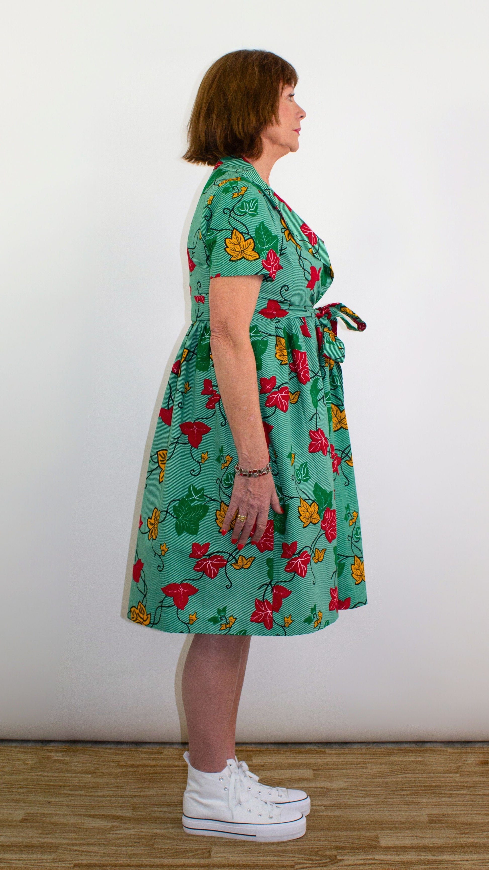 A side view of a model posing in a knee-length green print dress with leaf elements, highlighting the tie belt, tied into an elegant bow.
