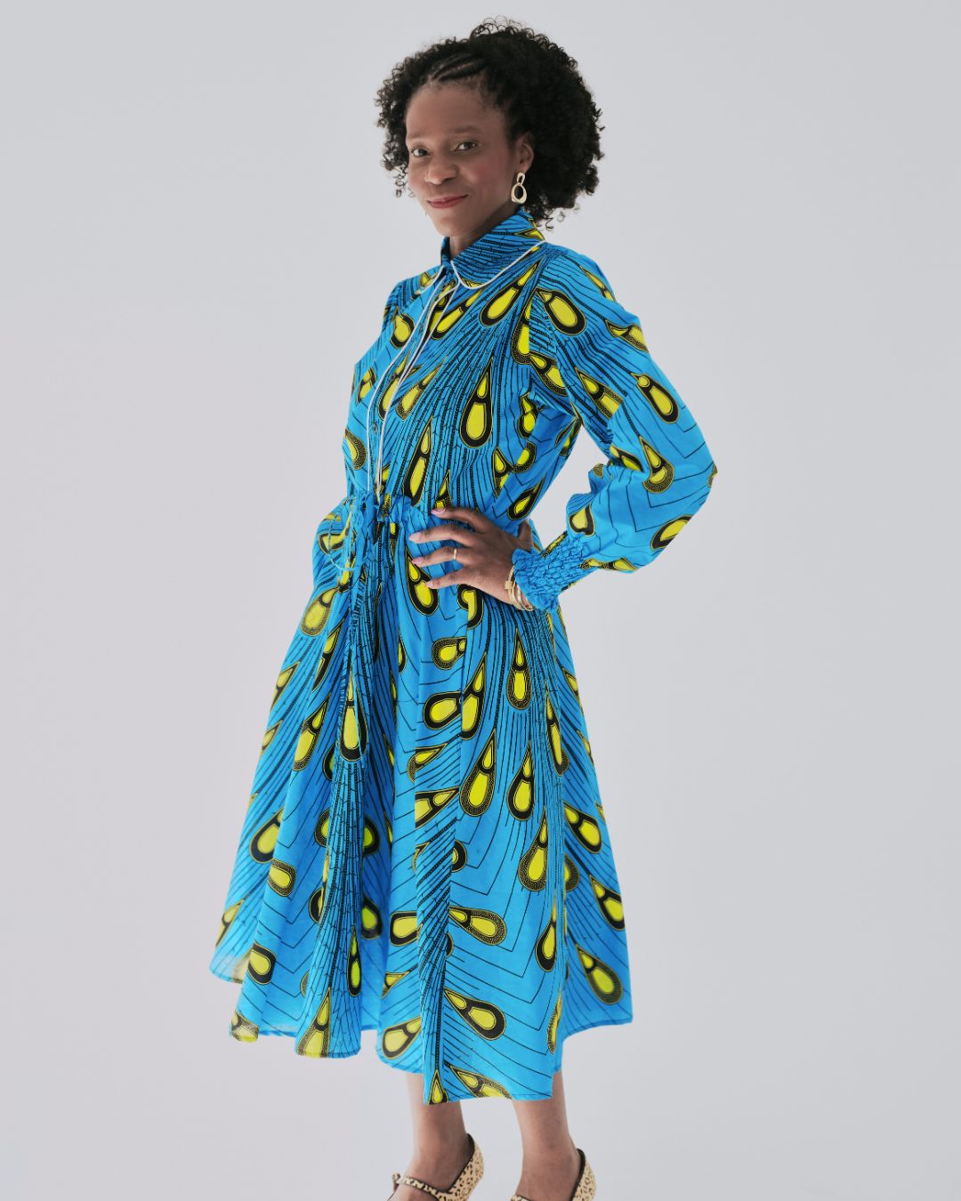 Model wearing a vibrant blue Ankara shirt dress with bold yellow teardrop pattern, full midi skirt, and long gathered sleeves. The dress features a classic collar and adjustable waist tie, styled for a flattering silhouette.