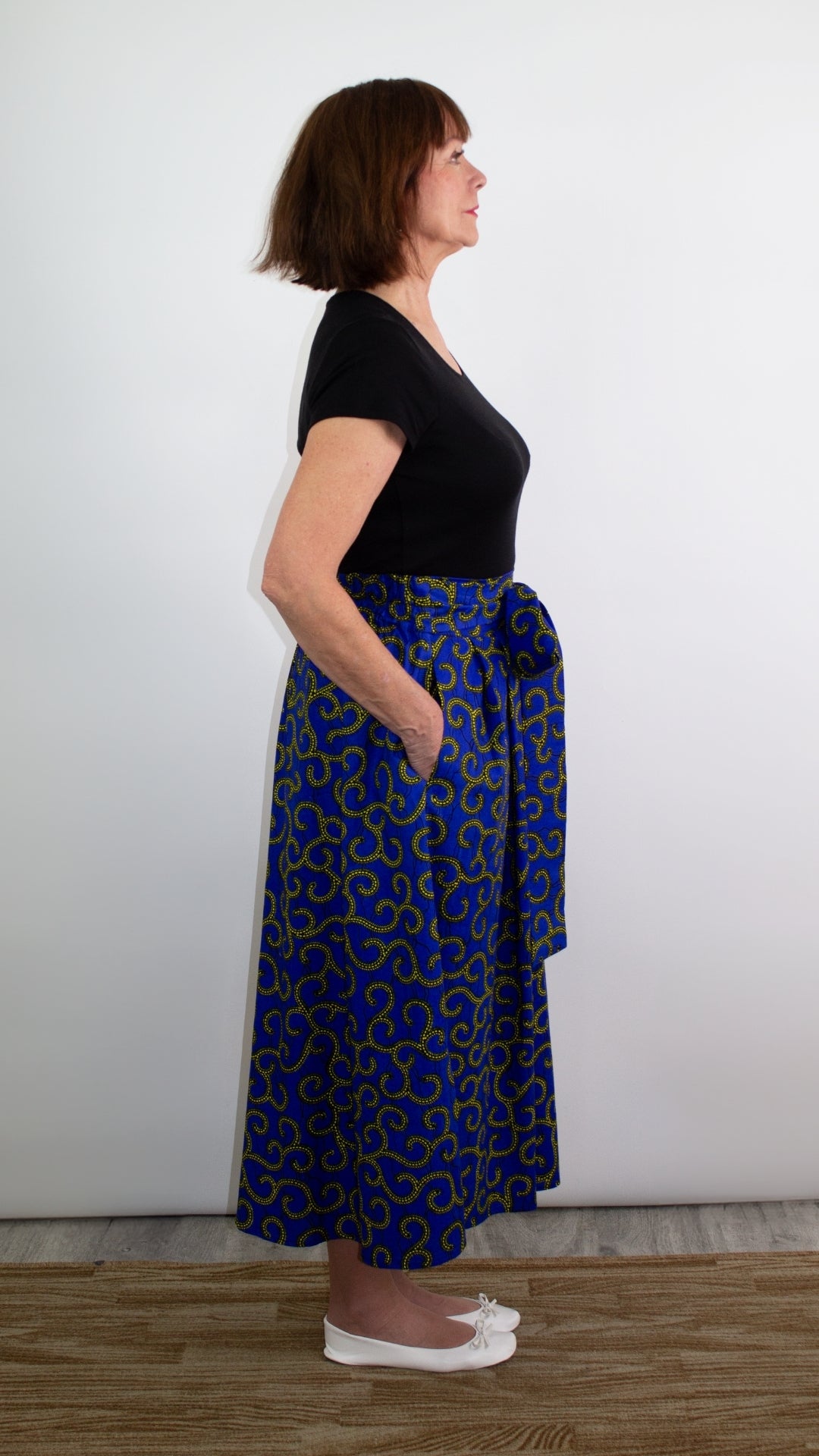 Side of model posing in a swirly blue skirt with golden elements showcasing comfortable deep pockets and a beautiful tie belt in a bow.