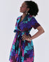Woman wearing a colorful fan-leaf print wrap dress in fuchsia, teal, purple, and black, featuring a notched collar, short sleeves, and a waist tie; styled with wedge heels.