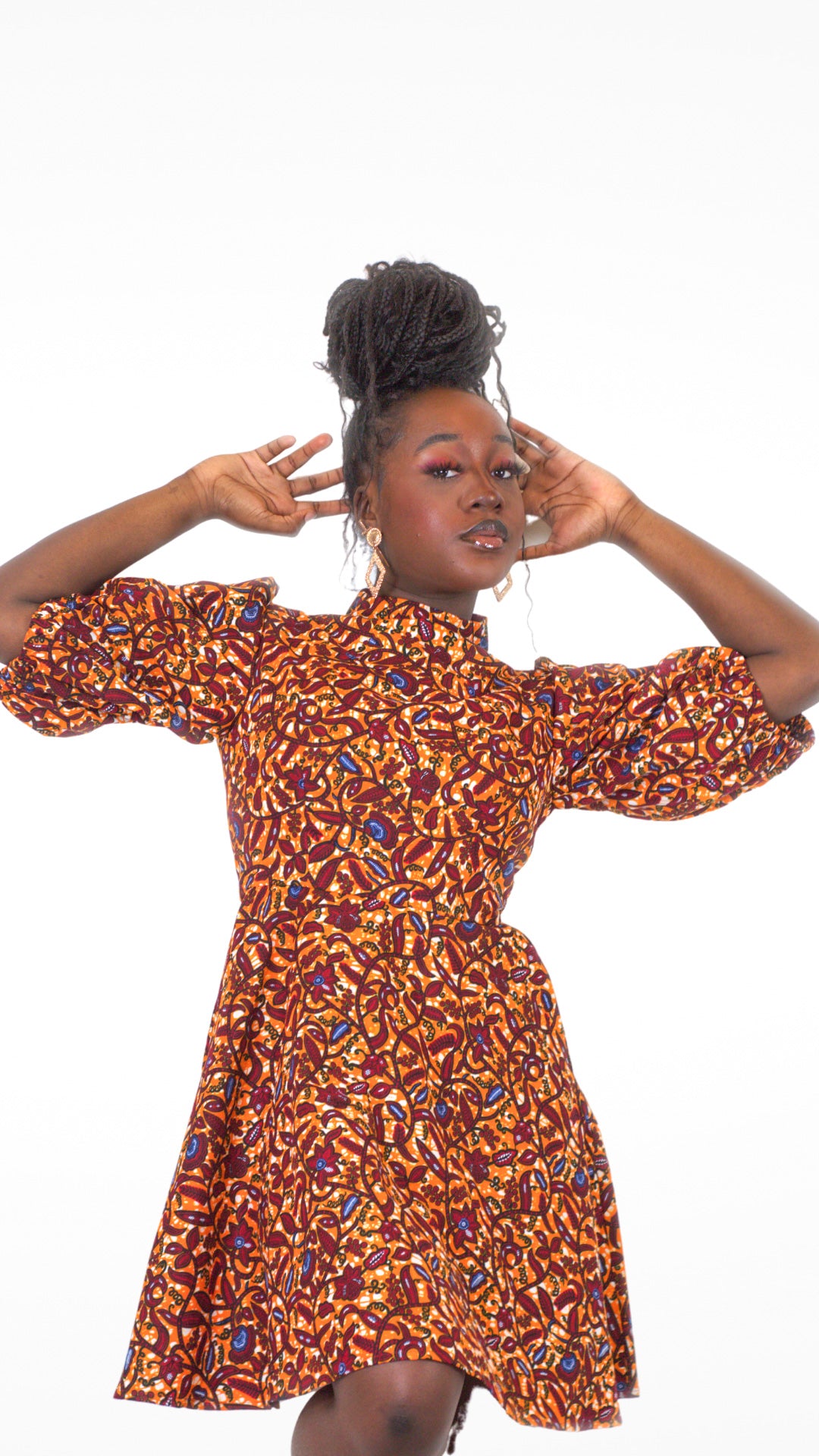 A woman confidently strikes a pose with arms up, showcasing the intricate elements of the neckline of her orange print puff sleeve dress.