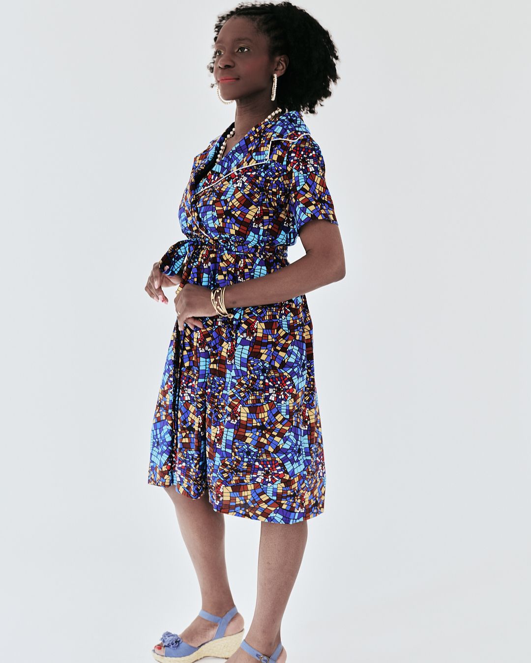 Woman in a knee-length, short-sleeved wrap dress with a geometric African print in blue, gold, and burgundy, styled with a waist tie and wedge heels.