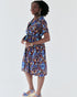 Woman in a knee-length, short-sleeved wrap dress with a geometric African print in blue, gold, and burgundy, styled with a waist tie and wedge heels.