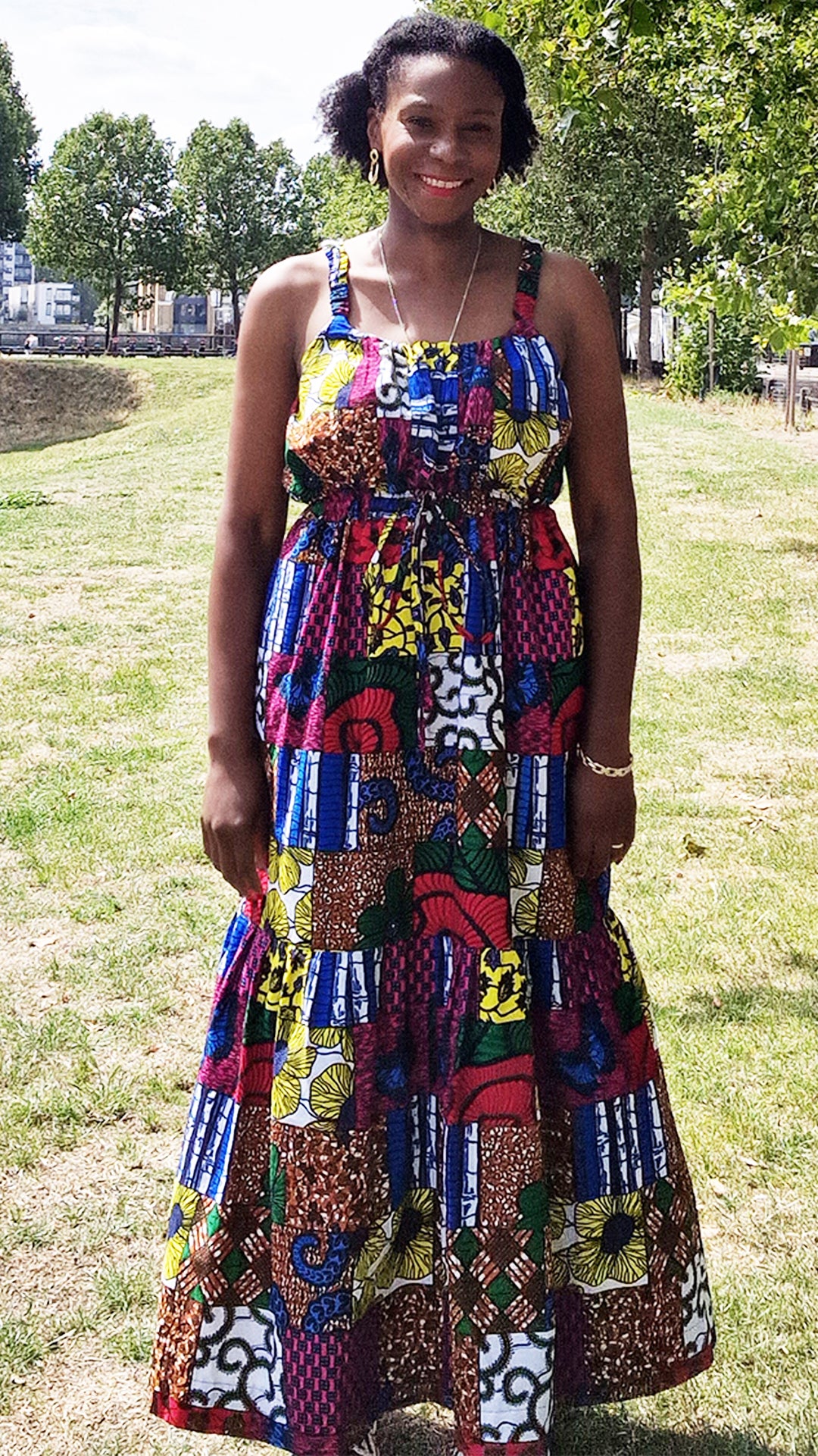 A woman posing with a radiant smile in a maxi long print patchwork dress, adorned with jewellery, in a park setting.